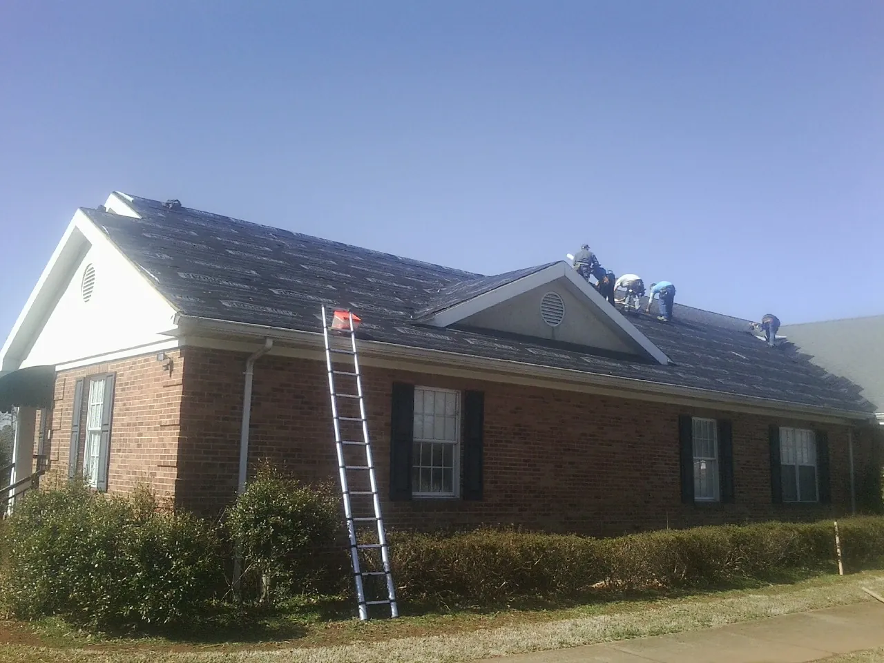 Crew tearing shingles off a brick ranch roof with a ladder set — Emergency Roof Repair work by our crew in Port Henry