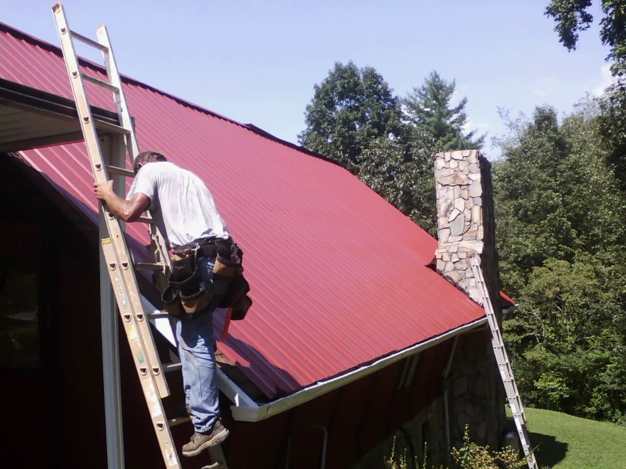 Roofer on a ladder beside a red metal roof and stone chimney — Soffit & Fascia Repair by Roofing Brothers in Port Henry
